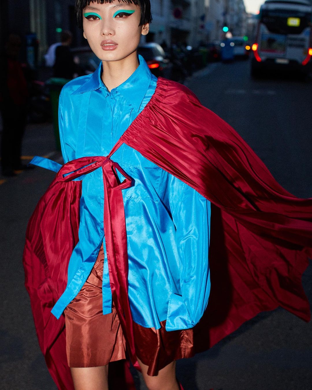 A young adult woman with teal makeup, wearing a shiny turquoise shirt and burgundy cape, stands on a city street with cars and buildings.