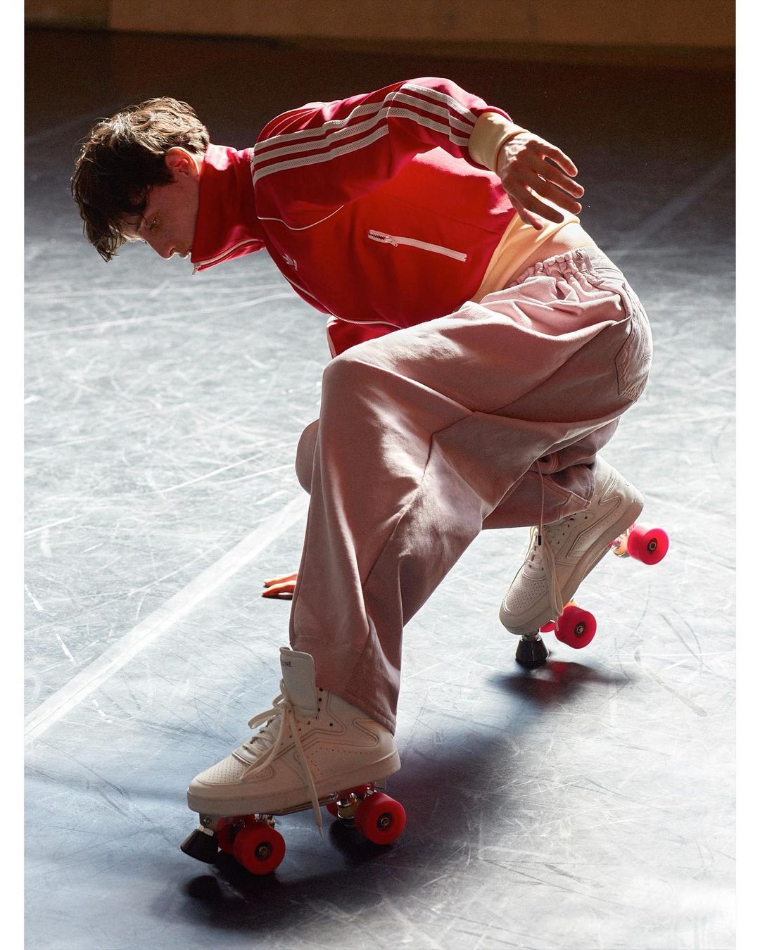 Young adult roller skating indoors wearing a red jacket, pink pants, and vintage-style roller skates with red wheels on a shiny floor.