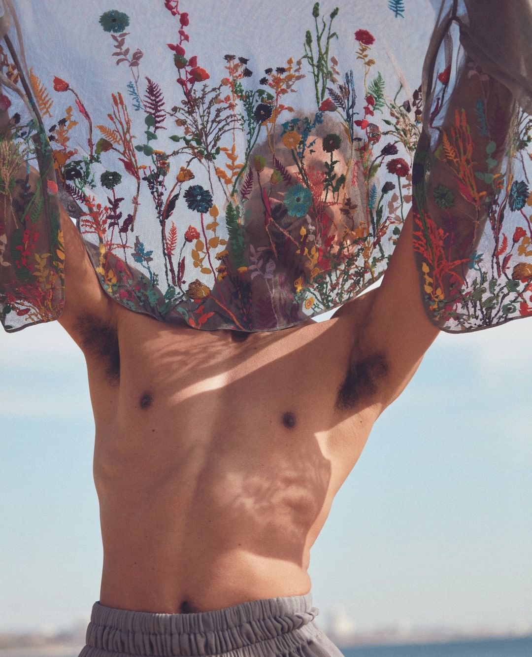 Man with grey sweatpants stands outdoors, shirtless, holding up a sheer, flower-embroidered textile against a light blue sky background.