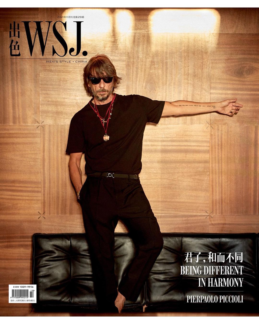 Pierpaolo Piccioli poses for a magazine cover. He is wearing a black t-shirt, black pants, and sunglasses in front of a wood wall.