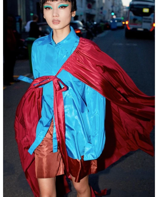 Fashionable person in a teal top and short copper skirt, styled with magenta fabric, poses on a city street with blurred cars.