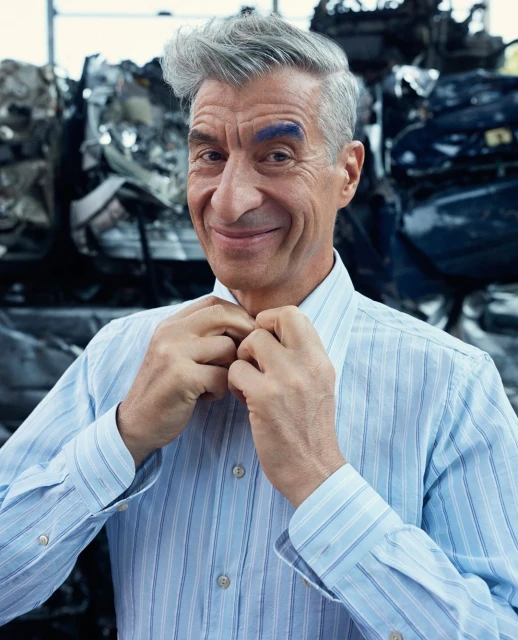 Close-up of a smiling older man with grey hair and blue eyebrow, wearing a striped button-down shirt against a blurry, dark background.