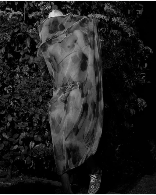 Monochrome image of a human figure draped in patterned translucent fabric, shoes visible, set against a leafy green backdrop.