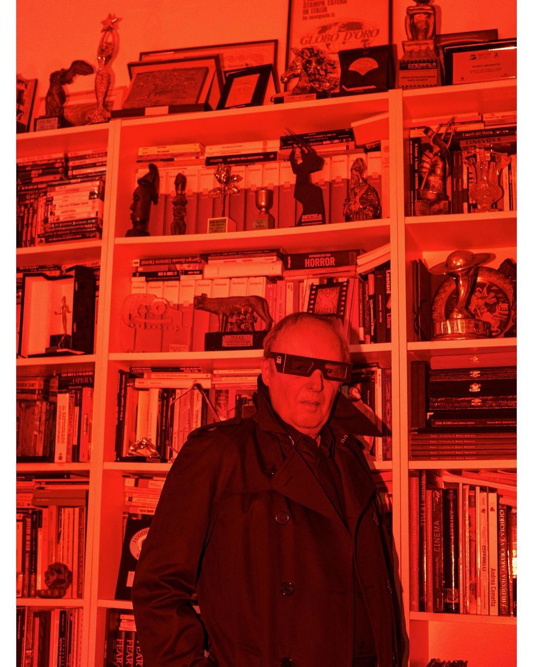 Man wearing 3D glasses, trench coat, standing in front of a bookshelf filled with books and trophies in a room with red lighting.