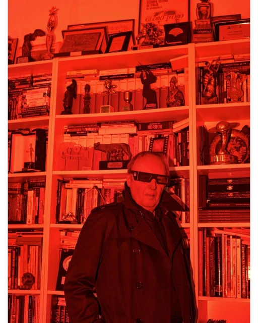 Man wearing 3D glasses, trench coat, standing in front of a bookshelf filled with books and trophies in a room with red lighting.