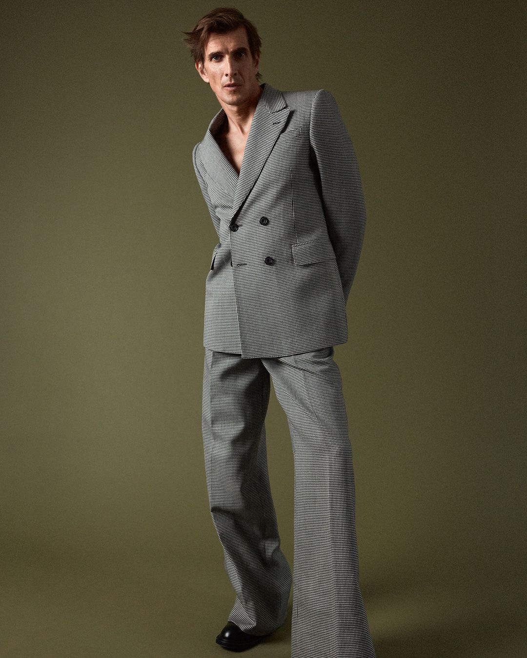 Man in a grey patterned double breasted suit posing against a solid olive green studio backdrop by Daniel Riera