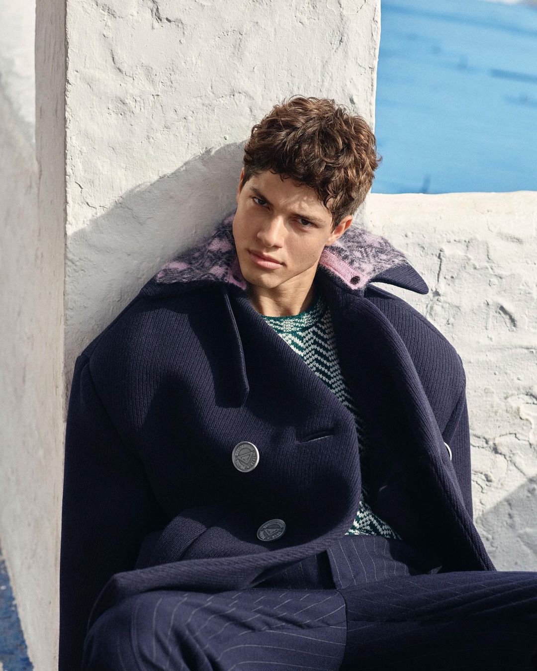 A young adult male sits outdoors wearing a navy coat with large buttons, a patterned sweater, and striped navy pants, against a stucco wall.