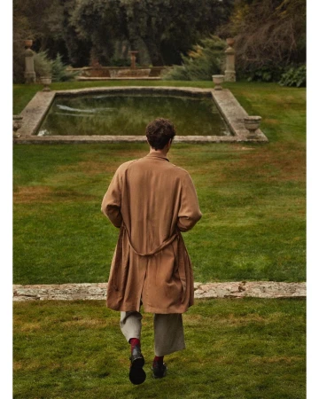 Back view of a person in a brown coat walking toward a pond in a green field surrounded by trees and stone architecture.