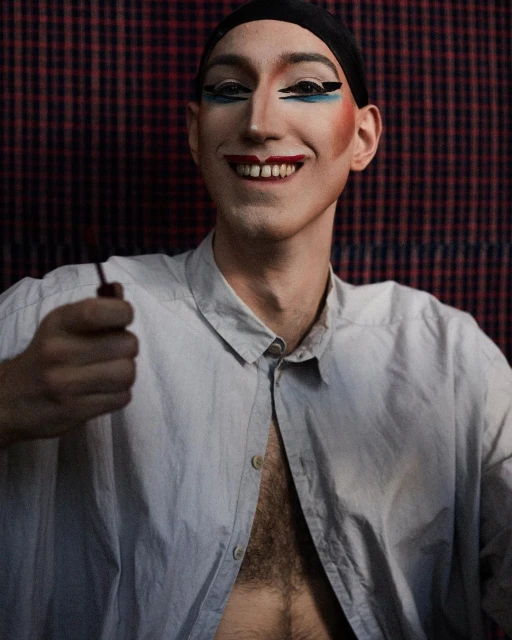 Smiling person with dramatic makeup in an unbuttoned shirt holding a cosmetic tool in front of a red plaid backdrop.
