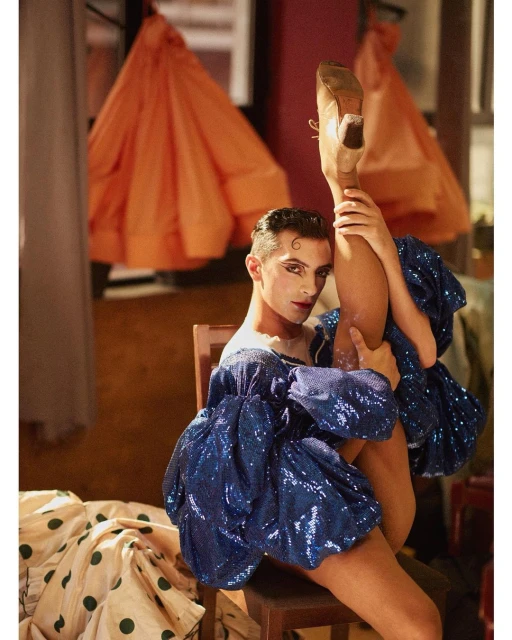 Man in blue sequin dress and heels poses on a chair, leg raised high, dramatic makeup, orange dresses hanging in the background.