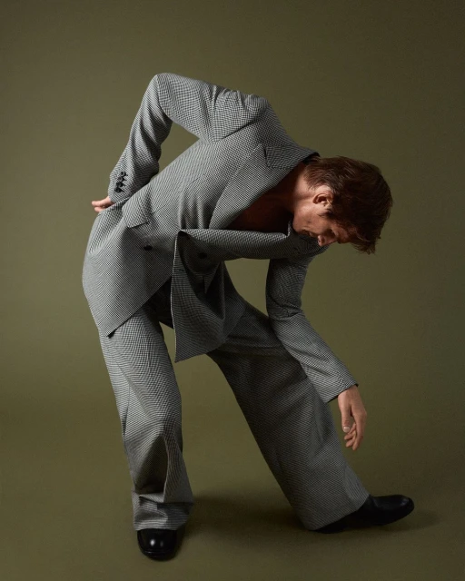 Young man in a grey houndstooth suit, bent over in an artistic pose on an olive-green background, fashion forward androgynous style.