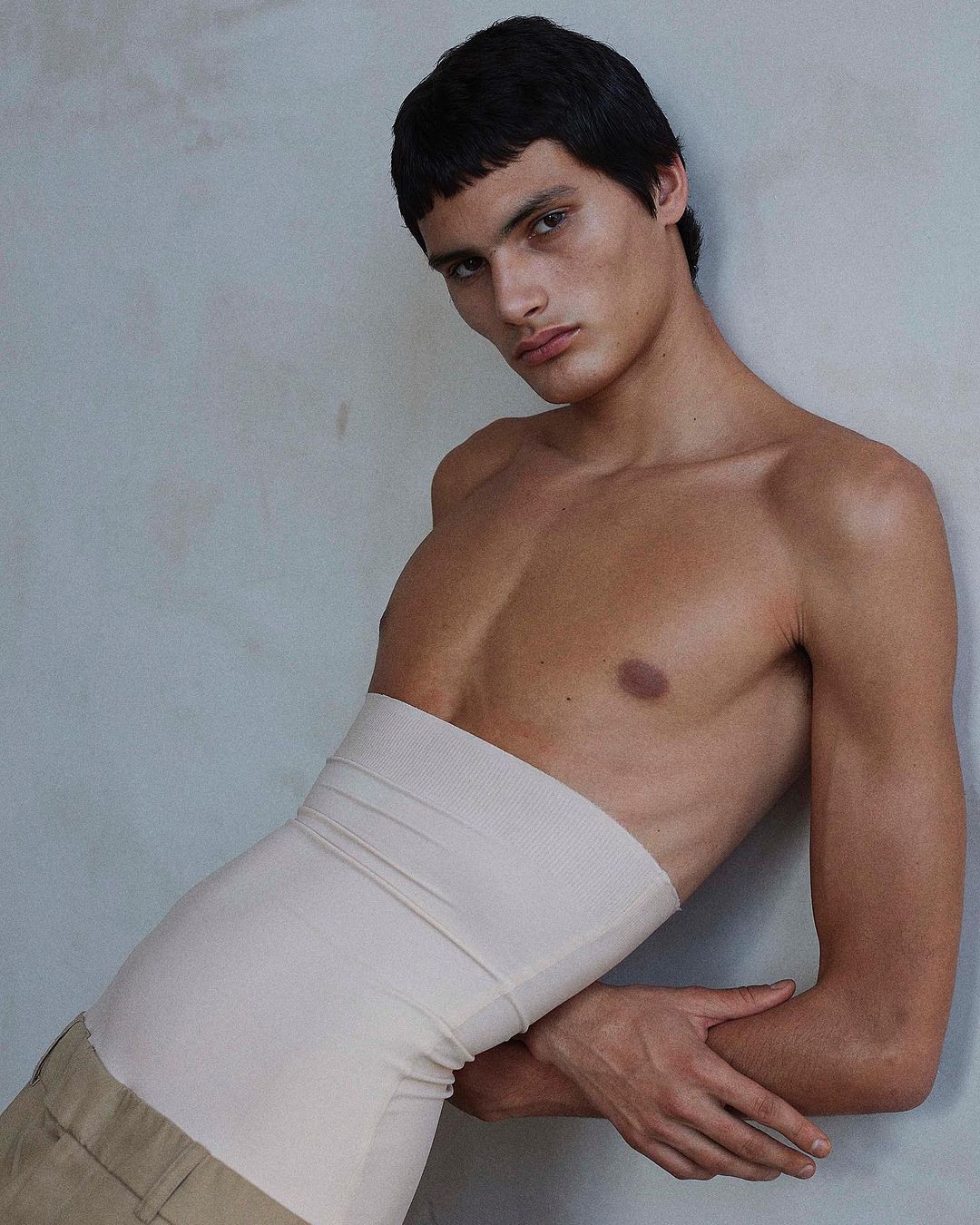 Portrait of a shirtless adult male fashion model posing with a neutral expression against a neutral gray wall wearing a tan skirt.