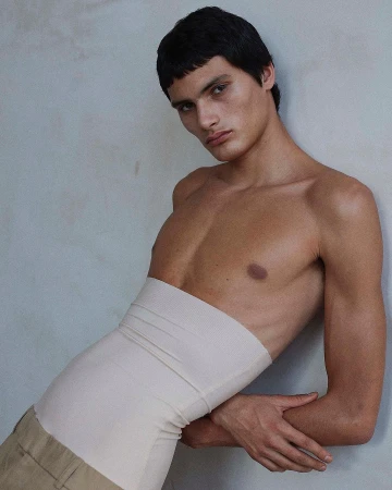 Shirtless male model wearing a high beige waist wrap leaning against a muted textured wall in Paris by photographer Daniel Riera.