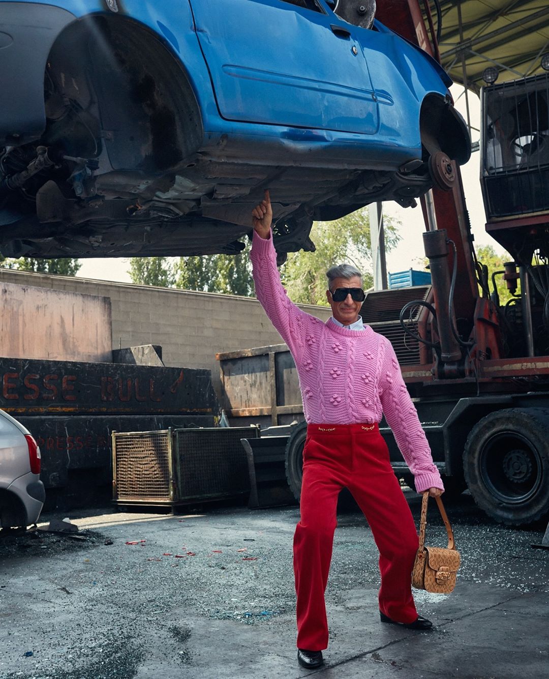 A man in a pink sweater stands under a blue car held aloft by a crane in a junkyard, holding a tan purse with sunglasses and red pants.