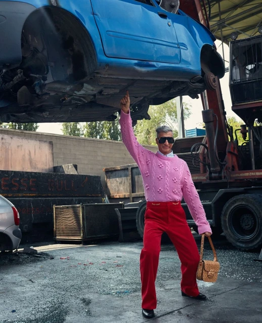 A man in a pink sweater stands under a blue car held aloft by a crane in a junkyard, holding a tan purse with sunglasses and red pants.