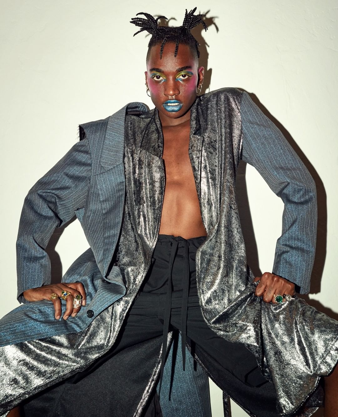 Person with creative makeup and hairstyle poses in the studio wearing a grey and silver blazer with a black top and unique expression.