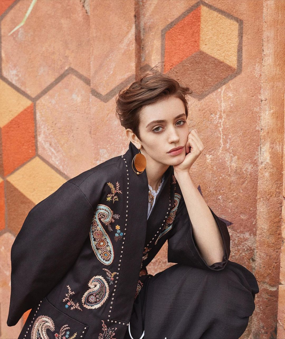 A woman with short brown hair poses in an embroidered kimono-style jacket against a wall painted with geometric designs.