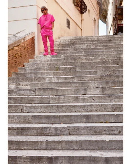 Adult male in a hot pink matching outfit stands on weathered stone steps outdoors under natural light in an urban setting.