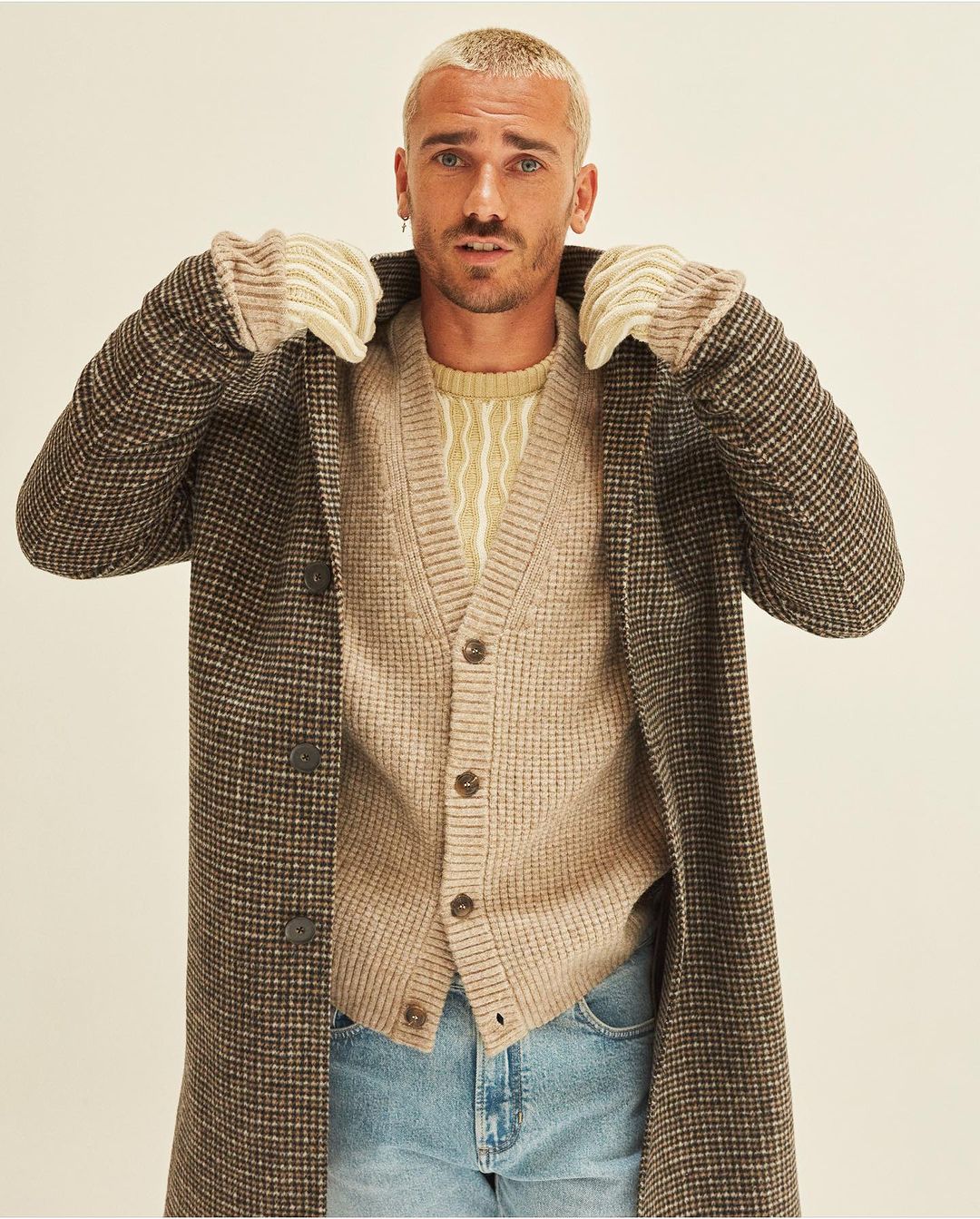 Portrait of adult male with blond hair, wearing brown plaid coat, beige cardigan, and light blue jeans in a neutral studio setting.