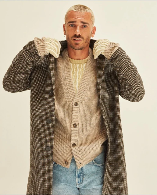 Portrait of adult male with blond hair, wearing brown plaid coat, beige cardigan, and light blue jeans in a neutral studio setting.