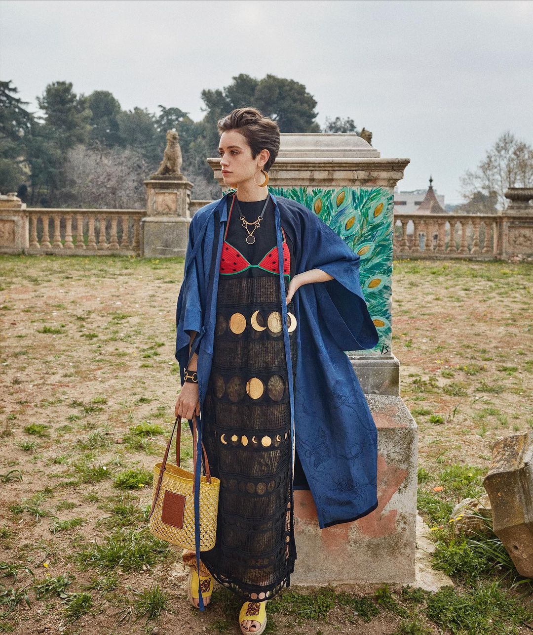 Woman in a black dress with moon phases, blue jacket, and straw bag poses in a park setting with statues and greenery. Fashion shot.
