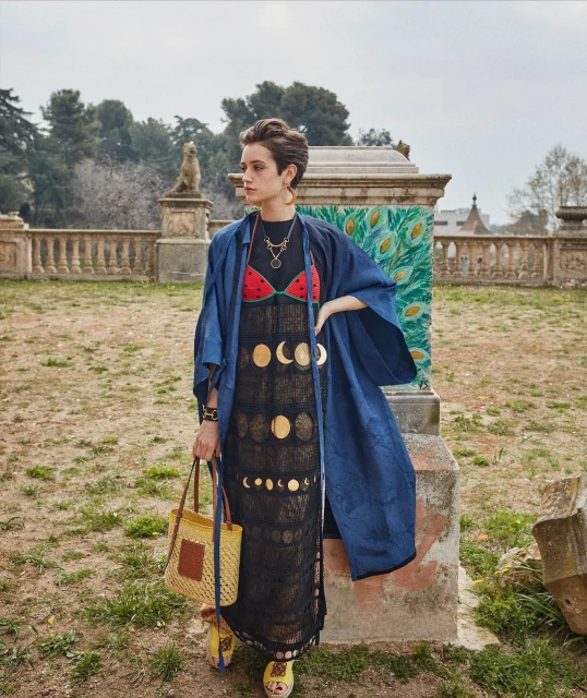 Woman in a black dress with moon phases, blue jacket, and straw bag poses in a park setting with statues and greenery. Fashion shot.