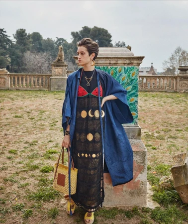 Woman in a black dress with moon phases, blue jacket, and straw bag poses in a park setting with statues and greenery. Fashion shot.
