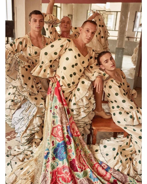 Group of four people wearing polka-dot dresses and decorative shawls posing indoors near a window and chair in a light-filled setting.