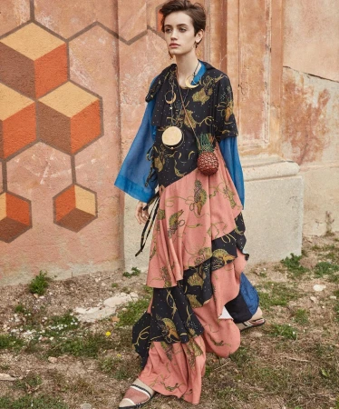 Model in patterned dark dress and blue sleeves posing outdoors against an orange wall with geometric painted patterns