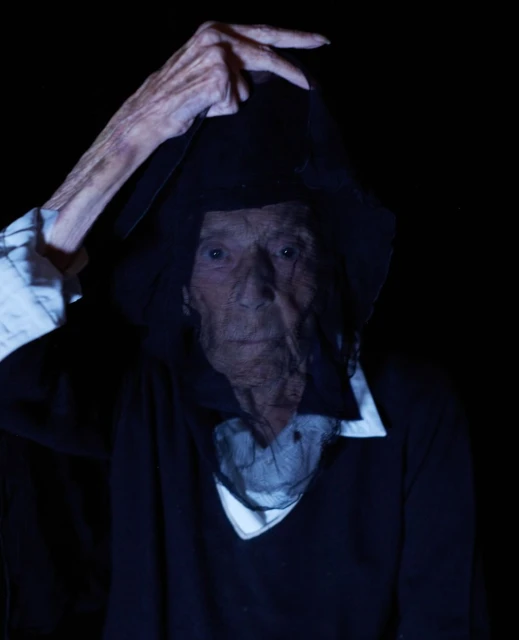 Eerie portrait of an elderly person with aged skin, hooded in dark fabrics, set against a stark black background, creating a dramatic visual.