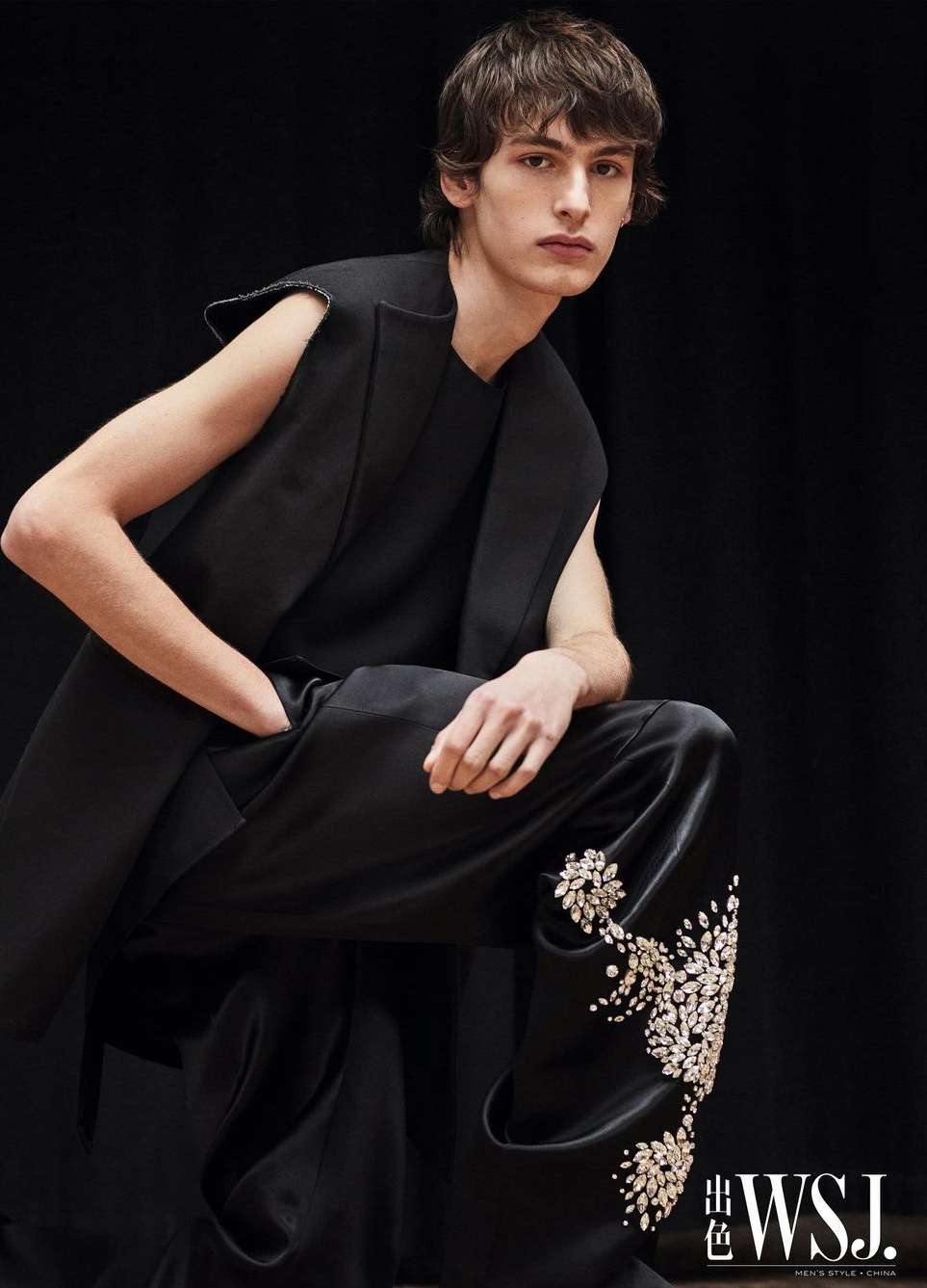 A light-skinned male model in a black sleeveless top and embellished pants, poses against a dark backdrop in a fashion shot.