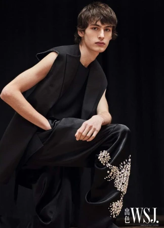 A light-skinned male model in a black sleeveless top and embellished pants, poses against a dark backdrop in a fashion shot.