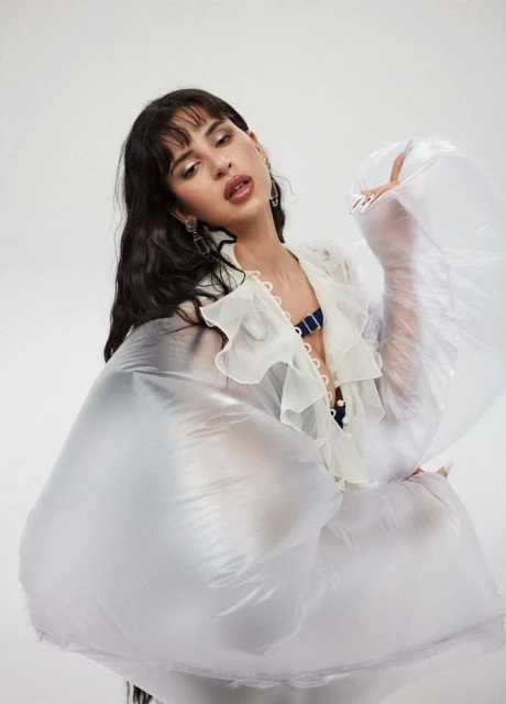 Portrait of a young woman in a ruffled blouse and puffed transparent sleeves against a white background, looking up.
