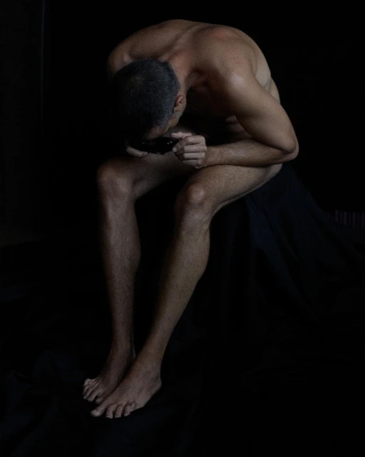 Nude man sits with head down in dark studio, emphasizing shadow and musculature in low-key lighting.