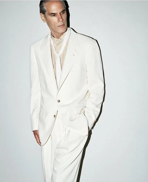 Mature man in an off-white suit stands in a photo studio. He is wearing an off-white suit with his hands tucked into his pockets.