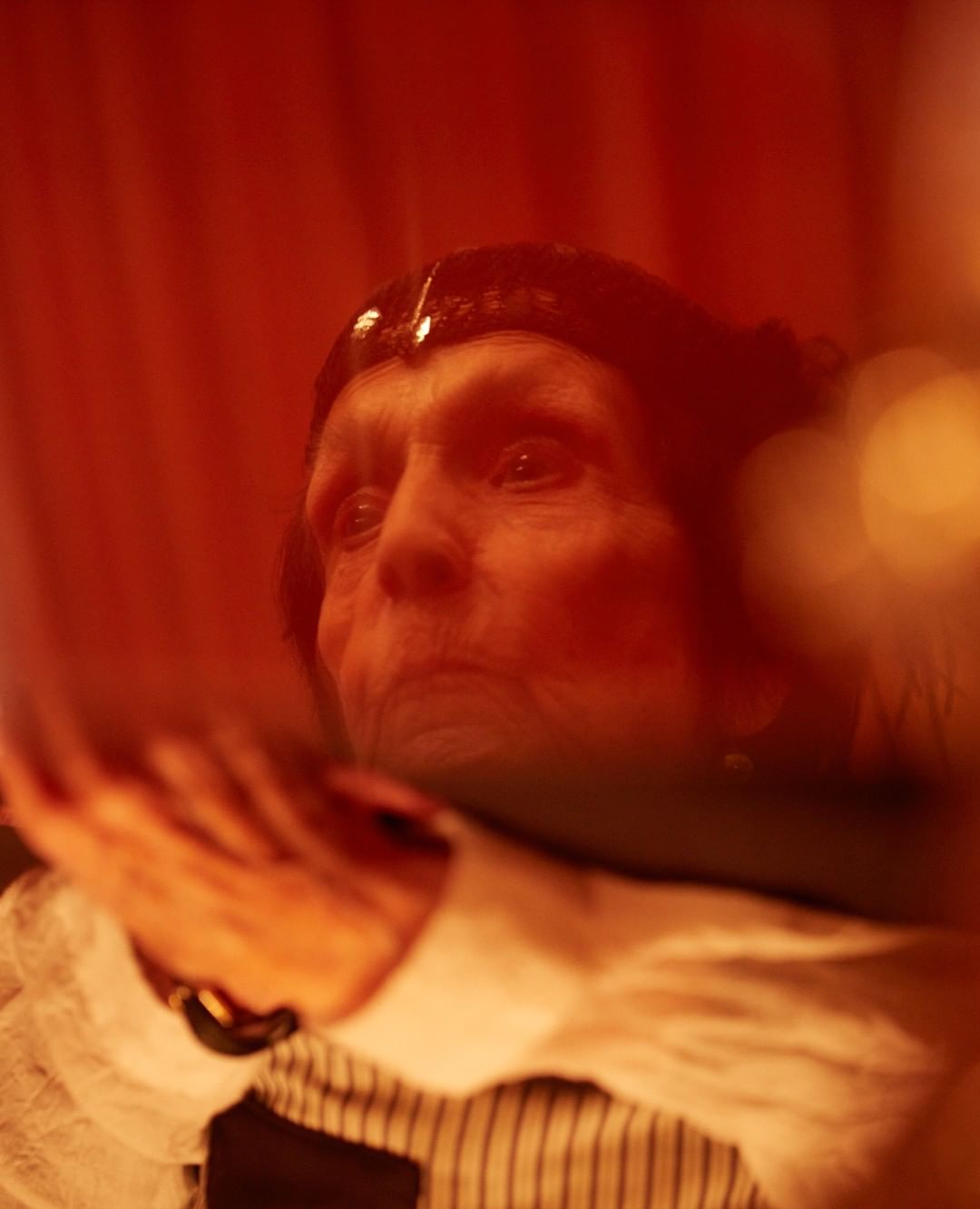 Close-up portrait of an elderly woman with a dark headband, wearing a white shirt with pinstripes, against a blurred orange background.