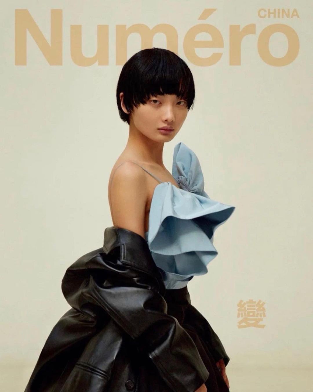 Model with short black hair wearing a light blue ruffled top and black leather jacket against a neutral backdrop.