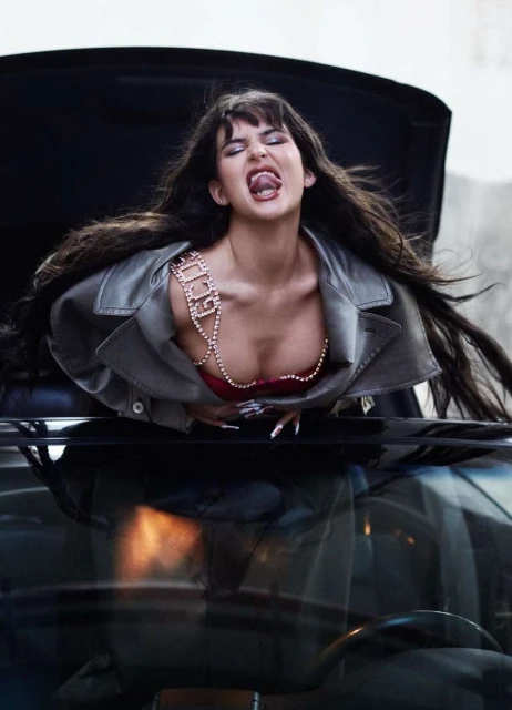 Stylish adult female model with long brown hair, wearing a grey jacket and beaded top, sticks her tongue out while leaning over a car hood.