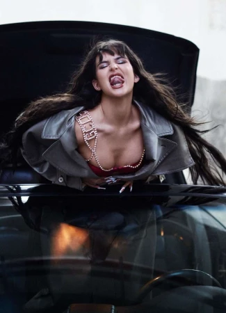 Stylish adult female model with long brown hair, wearing a grey jacket and beaded top, sticks her tongue out while leaning over a car hood.