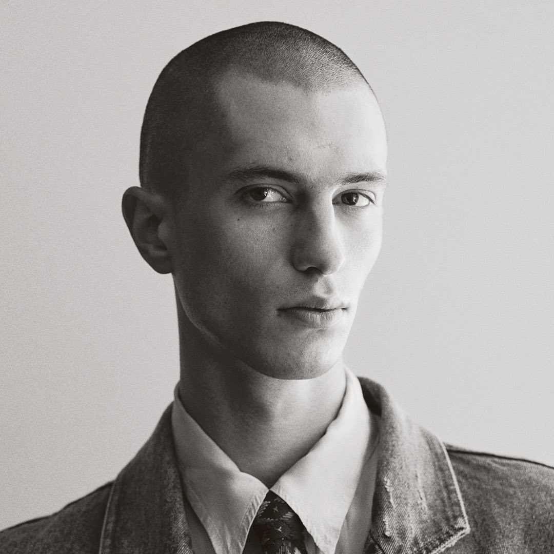 Monochrome portrait of a young adult male with a buzz cut, wearing a collared shirt, patterned tie, and denim jacket against a neutral background.