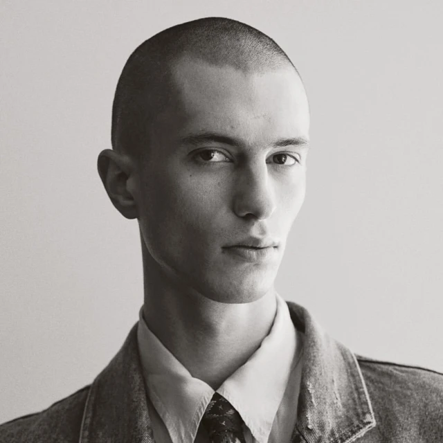 Monochrome portrait of a young adult male with a buzz cut, wearing a collared shirt, patterned tie, and denim jacket against a neutral background.