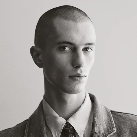Monochrome portrait of a young adult male with a buzz cut, wearing a collared shirt, patterned tie, and denim jacket against a neutral background.
