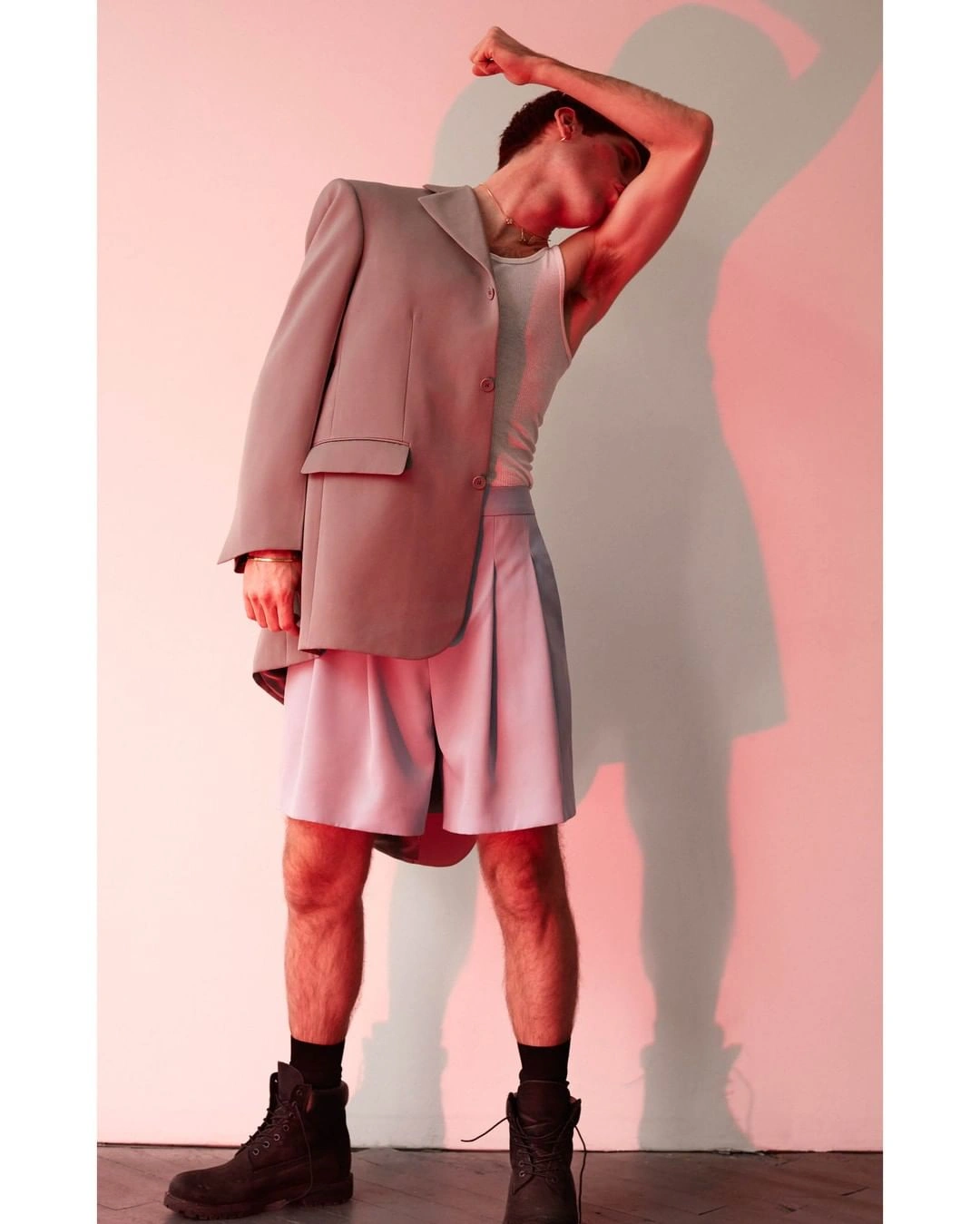 Man in oversized grey blazer, white tank top, light shorts and dark boots posing against a pink wall with shadow