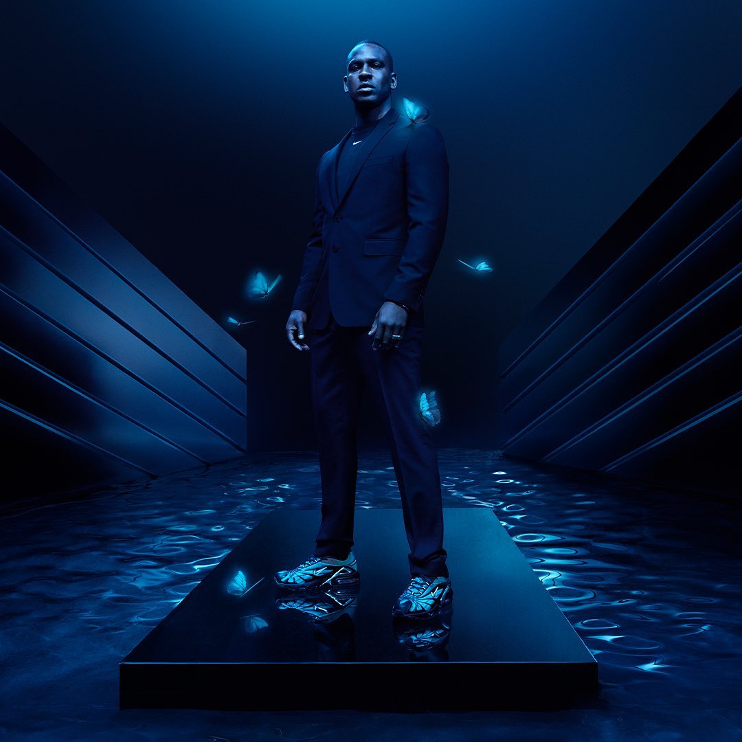 Stylish adult man in a dark suit stands on a platform in a blue-lit studio with artistic butterfly effects in a fashion photography shot.