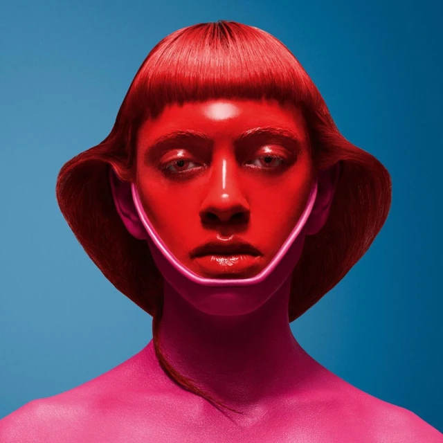 Close-up portrait of an adult woman with pink and red skin tones against a blue background, creating a vibrant and artistic visual.