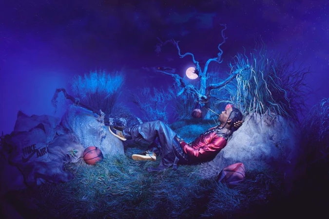Adult male poses while lying on grass next to basketballs and a Nike Air bag in a surreal nighttime outdoor scene with a moon.