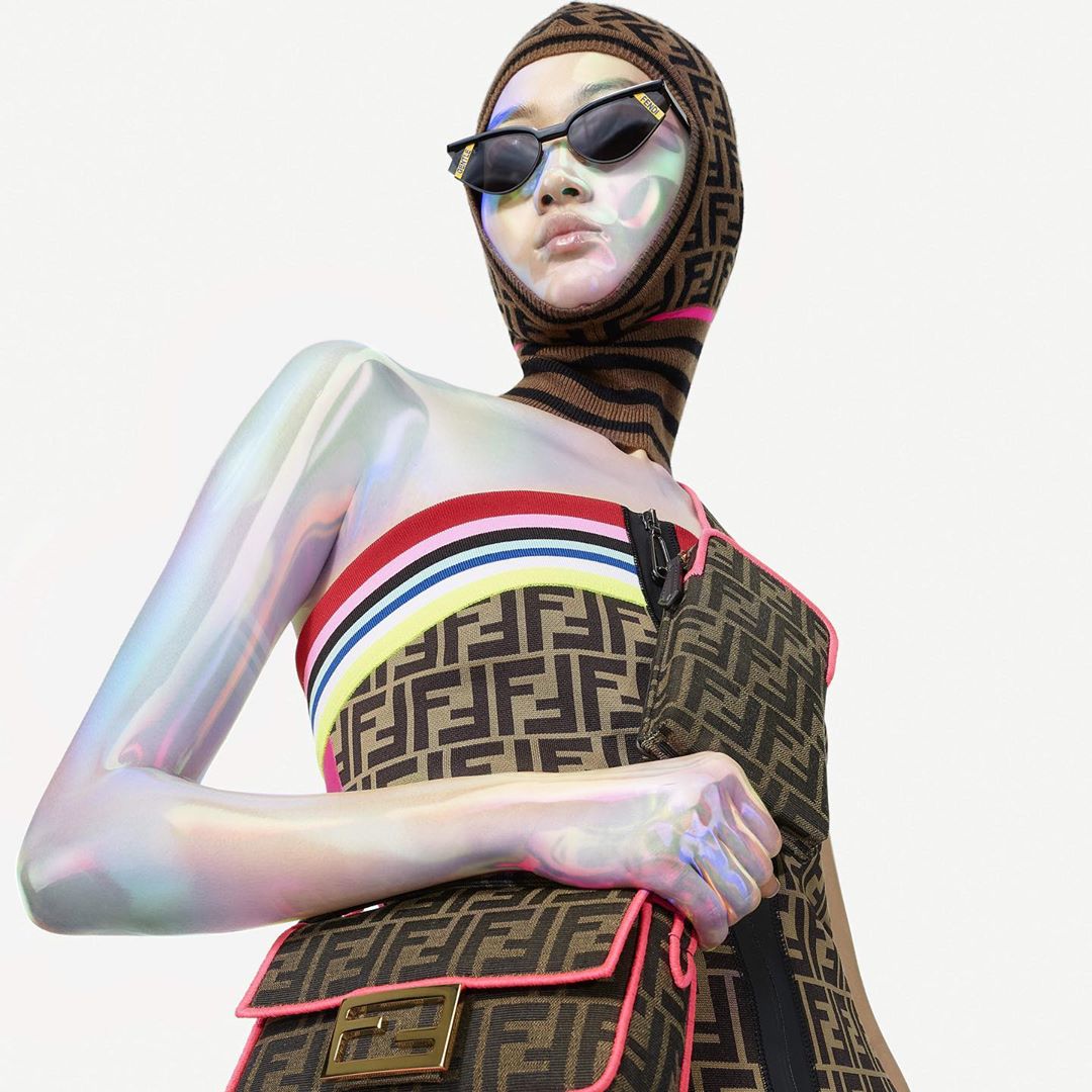 A woman models Fendi fashion wearing sunglasses, a patterned balaclava and dress, with a patterned handbag, against a white background.