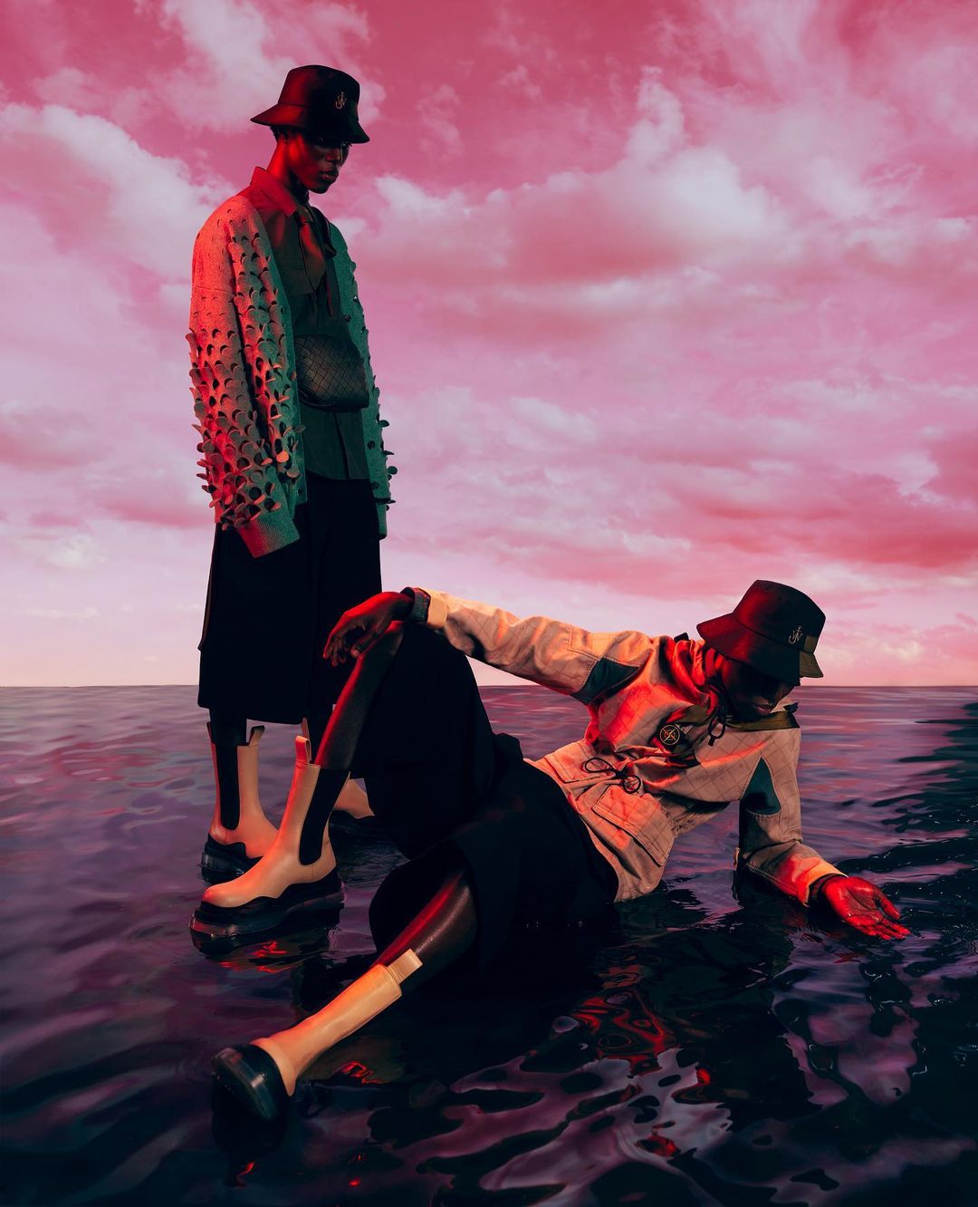 Two people wearing hats, jackets, and boots are posing in water under a pink, cloudy sky. One stands while the other reclines in the water.