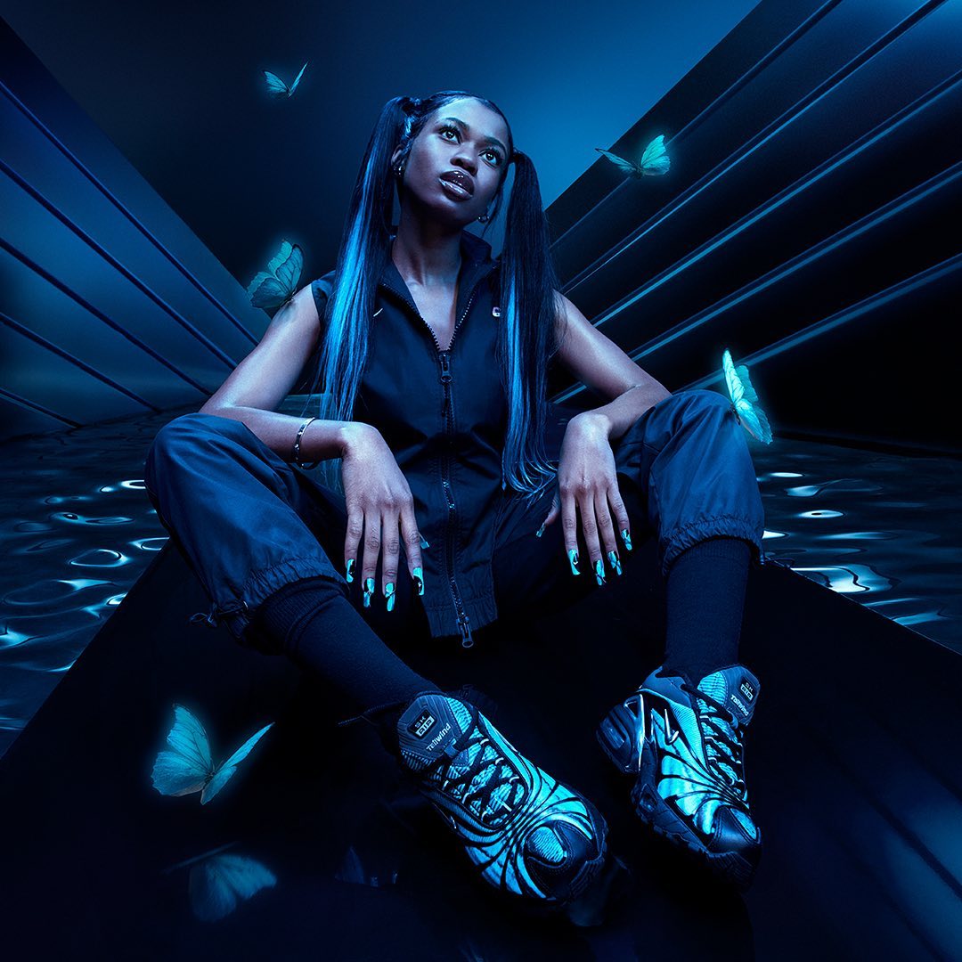 A Black female model with long hair, stylish nail art, and athletic shoes poses in a neon blue studio with glowing butterfly accents.