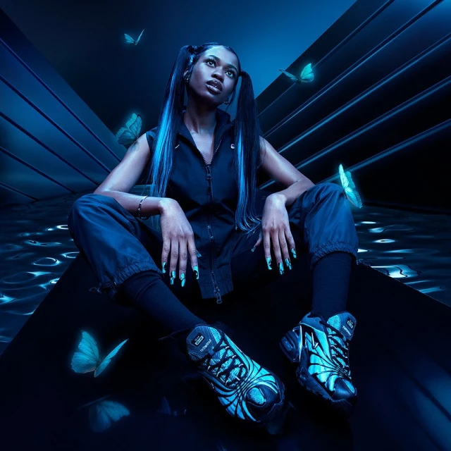 A Black female model with long hair, stylish nail art, and athletic shoes poses in a neon blue studio with glowing butterfly accents.